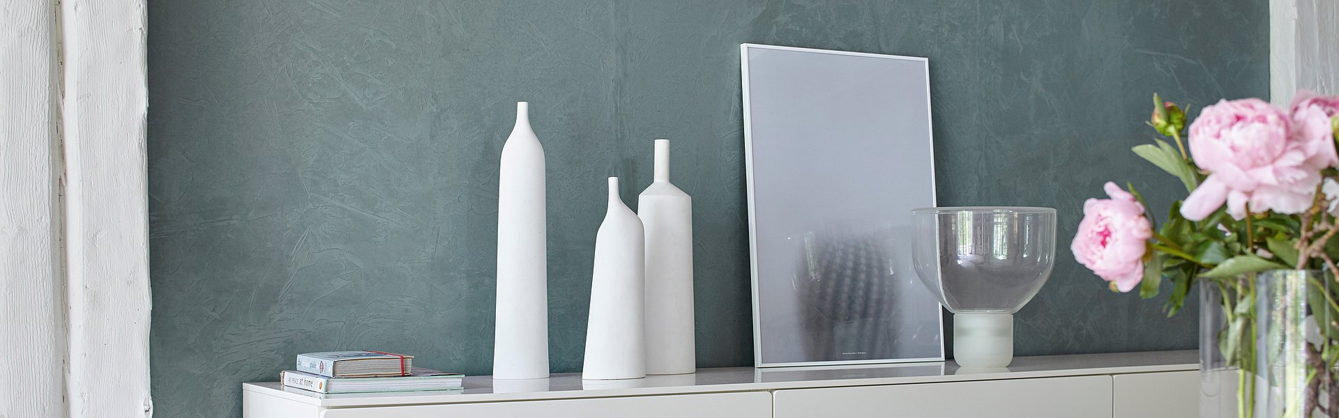 Sideboard an grüner Wand mit weißen Kerzen und roas Blumen als Deko